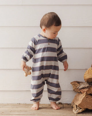 Toddler standing barefoot on a wooden deck near a stack of firewood, wearing a navy and cream striped long-sleeve romper with elastic cuffs, holding a wooden toy car.