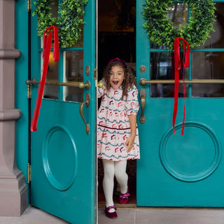 Girl wearing the Pink Chicken Maribelle Dress in Santa Garland print in a doorway hung with wreaths and red velvet bows