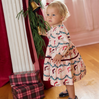 Baby wearing the Pink Chicken Maribelle Dress Set in pink Santa Garland print beside plaid-wrapped gifts
