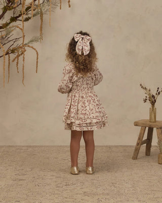 Back view of toddler girl in ivory floral toile dress with hair bow, standing on textured rug