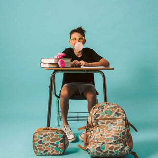 Boy blowing bubblegum at a school desk with matching Burlebo Bayside Camo backpack and lunchbox on the floor beside him