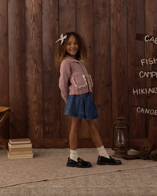 Girl with curly hair and a bow clip standing in profile, wearing a rosewood pink knit cardigan with scalloped collar and pockets, paired with a denim pleated skirt, white socks, and black loafers, set against a rustic wood-paneled backdrop with camp-themed signage.