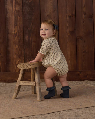 Toddler girl with a blue bow in her hair standing sideways at a small wooden stool, wearing a natural cream floral print bubble romper with ruffle collar and puff sleeves, paired with navy ribbed ankle socks, against a dark wood background.