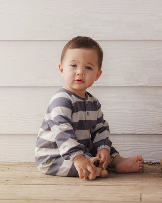 Baby sitting on a wooden porch wearing a navy and cream striped long-sleeve romper with wooden button closure, playing with a wooden toy car.