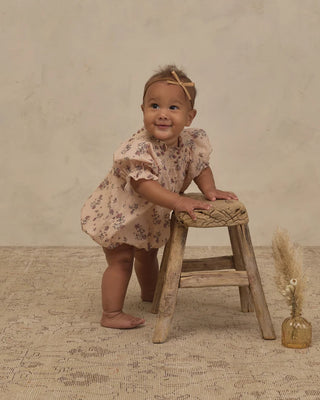 Baby girl in blush floral smocked romper standing at a wooden stool wearing a raffia bow headband