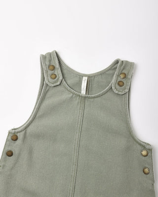 Close-up of a sage green twill overall dress showing the crossback shoulder straps with bronze snap buttons, round neckline, and side snap closures on the hip.
