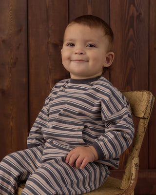 Toddler sitting in a vintage wooden chair against a dark wood-paneled background, wearing a striped navy, cream, and rust long-sleeve sweatshirt with front pocket and matching pants.