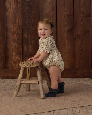 Toddler girl smiling at the camera while standing at a small wooden stool, wearing a natural cream floral print bubble romper with ruffle collar and puff sleeves, paired with navy ribbed ankle socks.