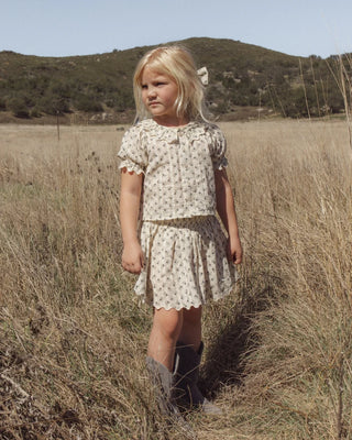 Girl standing in a golden grass field with hills in the background, wearing a matching ivory floral print top and scalloped-hem skirt set with a hair bow and cowboy boots.