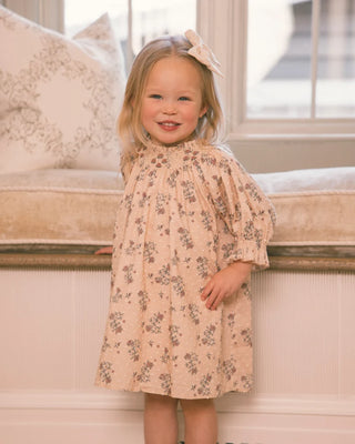 Toddler girl smiling in blush floral smocked dress with puff sleeves, standing by a window seat