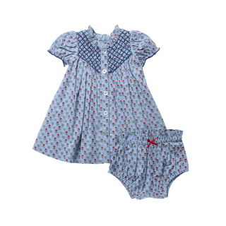 Product flat lay of a light blue carnation print dress and matching bloomer shorts, featuring diamond navy smocking at the bodice, puff sleeves, glitter button front closure, and a red satin bow accent on the bloomers, on a white background.