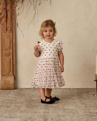 Young girl in a white dress with red patterns standing indoors against a beige wall.
