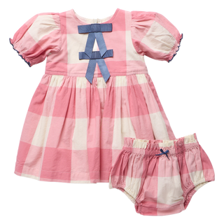 Product flat lay, front view, of a pink and cream check dress and matching bloomer shorts, featuring a navy grosgrain bow at the neckline and a smaller navy bow accent on the bloomers, on a white background.