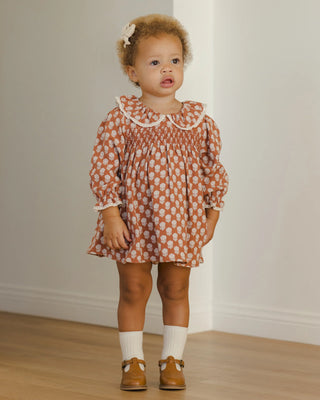 Young child wearing a patterned dress standing on a wooden floor.