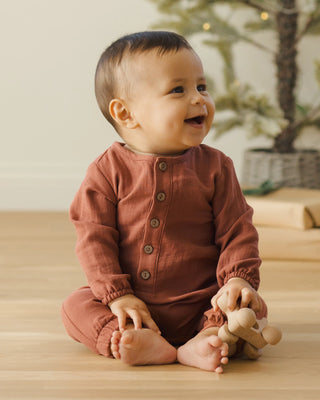 Baby in a rust-colored onesie sitting on a wooden floor with a toy.
