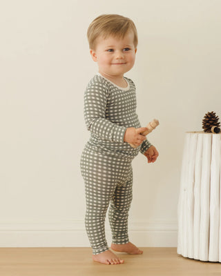 Child wearing a green and white checkered outfit standing on a wooden floor.