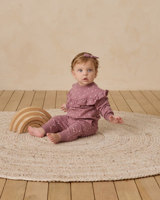 Baby in a pink outfit sitting on a round rug with a wooden rainbow toy.