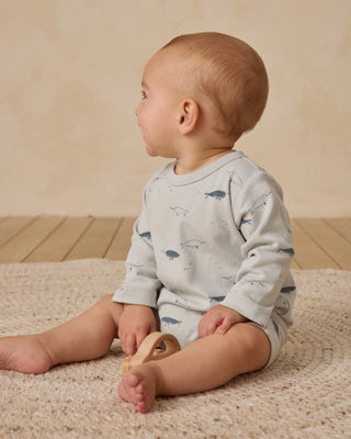 Baby sitting on a carpeted floor wearing a light blue onesie with animal prints.