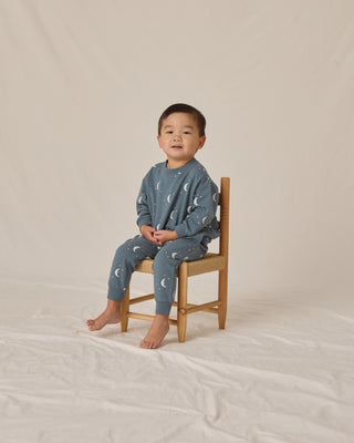 Child wearing blue pajamas with moon pattern sitting on a wooden chair against a beige background