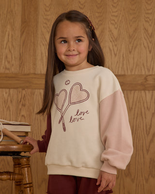 Young girl wearing a cream sweater with pink sleeves and a heart design, standing against a wooden paneled wall.