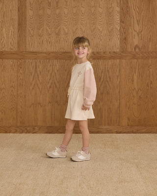 Young girl in a light pink dress standing against a wooden paneled wall.