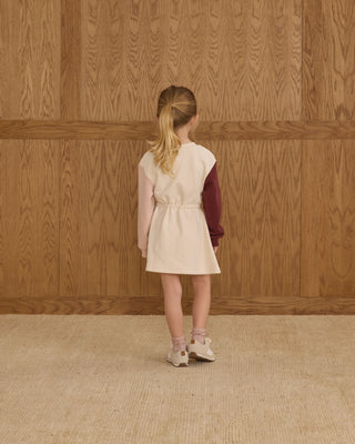 Young girl standing against a wooden paneled wall wearing a cream dress and maroon sweater.