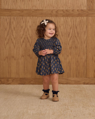 Young girl in a patterned dress standing against a wooden paneled wall.