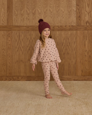Child wearing a polka dot outfit with a matching hat against a wooden paneled wall.