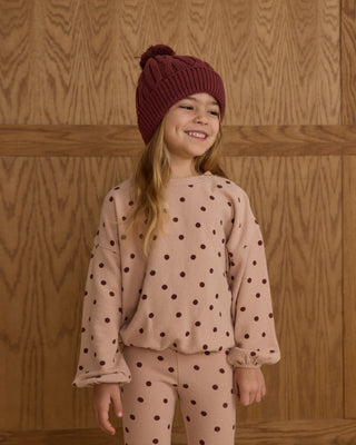 Young girl wearing a polka dot outfit with a maroon knit hat against a wooden background