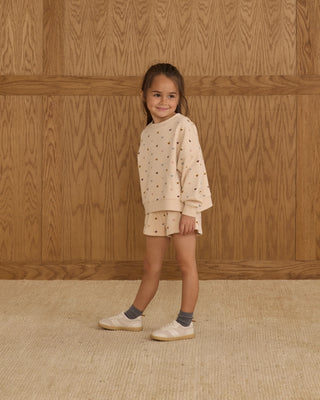 Young girl wearing a beige outfit with small patterns against a wooden paneled wall.