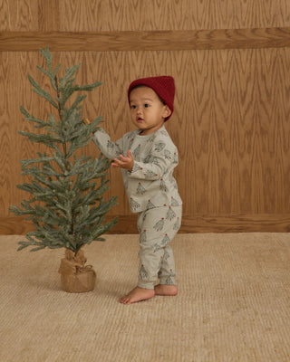 Child in a red hat standing next to a small tree against a wooden wall.