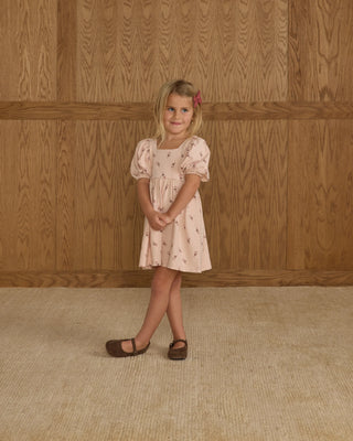 Young girl in a floral dress standing against a wooden paneled wall.