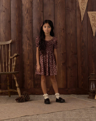 Girl with a floral hair clip standing against a rustic wood-paneled backdrop with camp-themed banners, wearing a mulberry floral print dress with ruffled square neckline and puff sleeves, paired with white ruffle socks and black loafers.