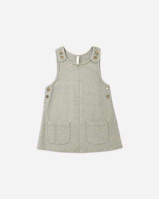 Sage green cotton overall dress laid flat, featuring a center front seam, two front patch pockets, and bronze snap buttons at the shoulder straps and side hips.