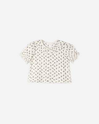 Ivory floral print top laid flat, featuring a ruffled collar, short puff sleeves with scalloped cuffs, and a front button placket.