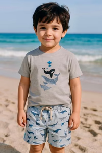 Boy wearing a gray t-shirt with whale designs and blue shorts with whale patterns on a beach.