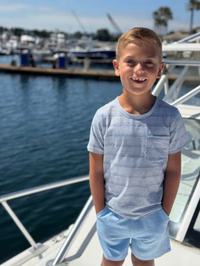 A young boy smiling on a boat deck, wearing a light blue and white striped short-sleeve t-shirt and matching light blue shorts, with water and marina boats in the background.