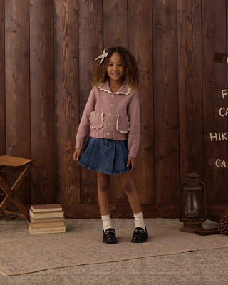 Girl with curly hair and a pinwheel-shaped hair clip smiling and standing against a rustic wood-paneled backdrop with camp signage, wearing a rosewood pink knit cardigan with scalloped collar and pockets, paired with a denim pleated skirt, white socks, and black loafers.
