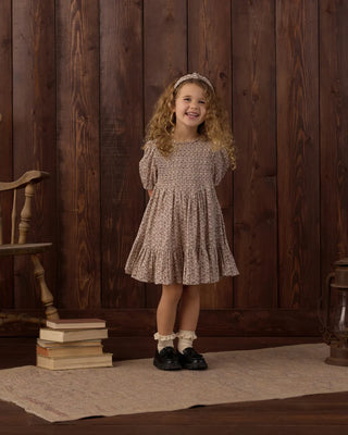 Girl with curly hair smiling and standing against a dark wood-paneled backdrop, wearing a matching floral print headband and a taupe floral smocked-bodice tiered dress, paired with white ruffle socks and black loafers.