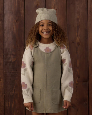 Smiling young girl with curly hair wearing a cream knit beanie, an ivory sweater with pink floral pattern, and a sage green overall dress with front patch pockets, standing against a dark wood plank background.