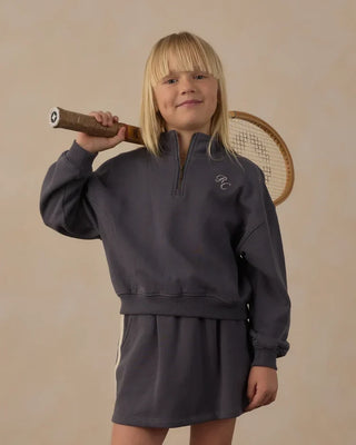 Close-up of a girl smiling at the camera, holding a vintage wooden tennis racket over her shoulder, wearing a midnight blue quarter-zip sweatshirt with embroidered 'RC' logo at the chest.