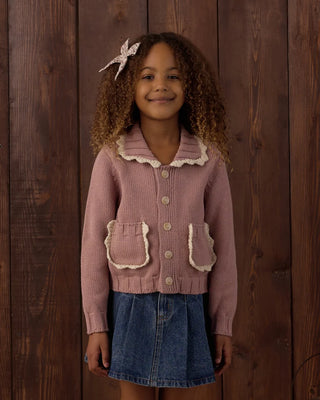 Girl smiling at the camera, wearing a rosewood pink knit cardigan with scalloped cream-trimmed collar and pockets, buttoned over a denim skirt, against a dark wood-paneled background.