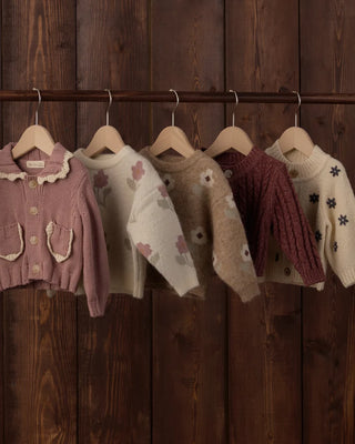 Five children's knit sweaters hanging on wooden hangers against a dark wood-paneled wall: a rosewood pink cardigan with scalloped collar, an ivory floral crewneck, a tan floral crewneck, a burgundy cable-knit cardigan, and an ivory sweater with dark floral embroidery.