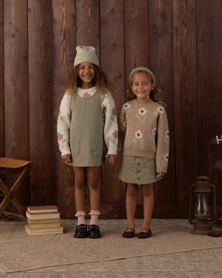 Two girls holding hands and smiling, one wearing a knit beanie with an ivory floral sweater under a sage green overall dress with white ruffle socks and loafers, the other wearing a beret with a tan floral sweater and sage green button-front skirt with brown flats, against a dark wood-paneled background with camp props.