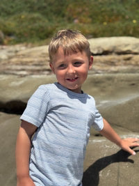 A young blonde boy smiling while wearing a light blue and white striped t-shirt, standing outdoors on a rocky beach with blurred green foliage in the background.